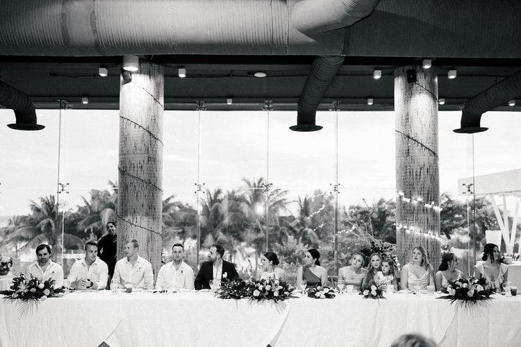 Wedding party seated at a long reception table beneath palm trees at dusk at a Playa del Carmen beachfront venue.