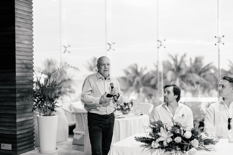 A guest gives a wedding toast into a microphone as groomsmen look on at an open-air reception with palm trees behind the glass wall.