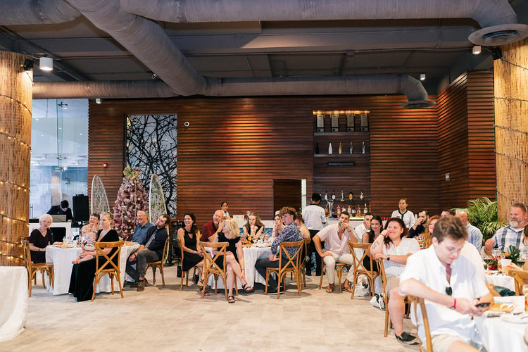 Wedding reception guests seated at round tables listen and laugh during toasts inside a wood-paneled venue with string lights.