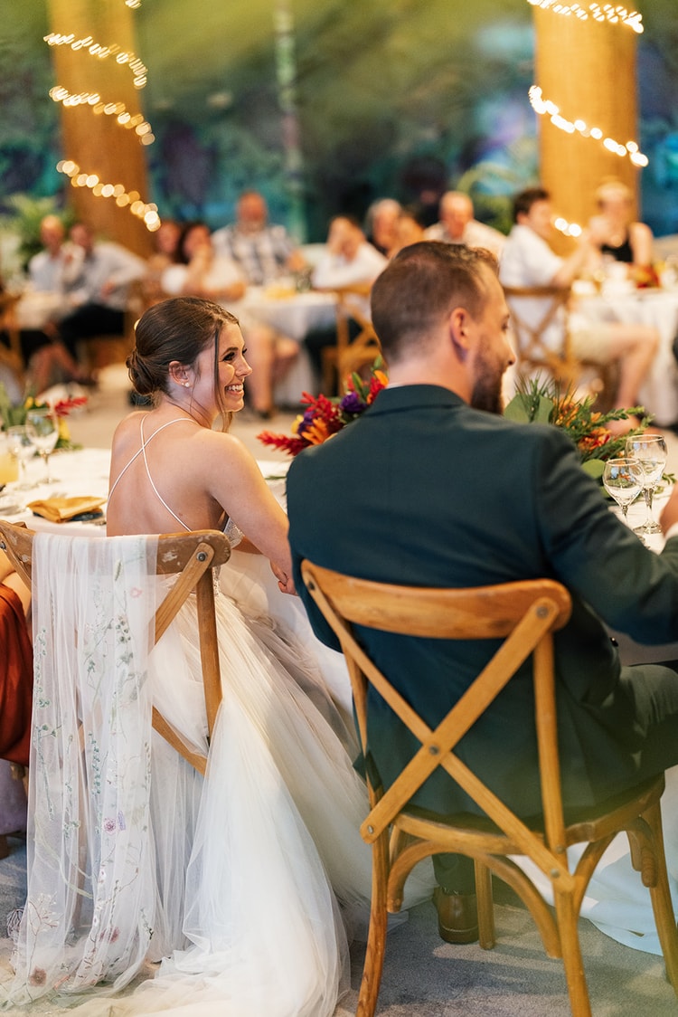 Bride laughs over her shoulder at the groom while seated at the reception table, her floral-embroidered veil draped over her chair.