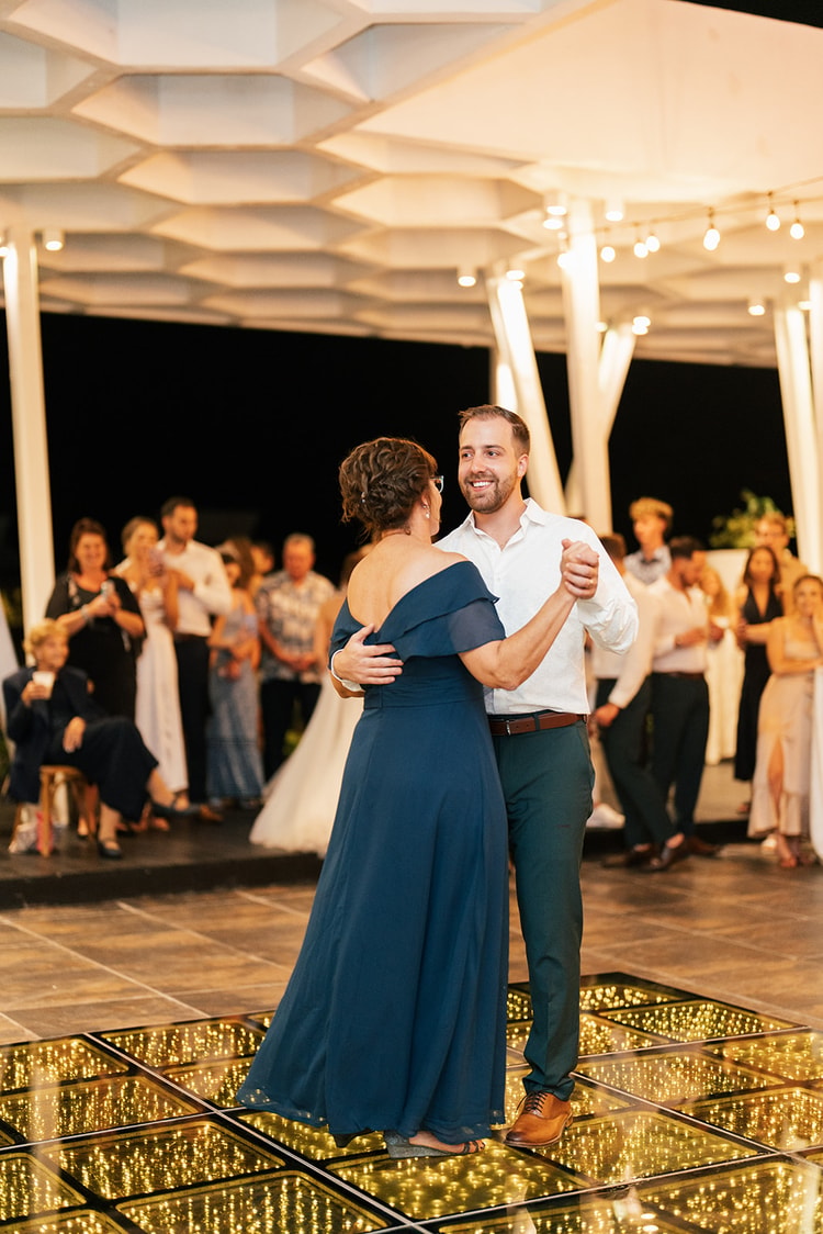 Groom dances with his mother in a navy off-shoulder gown as wedding guests watch from the edge of the illuminated dance floor.