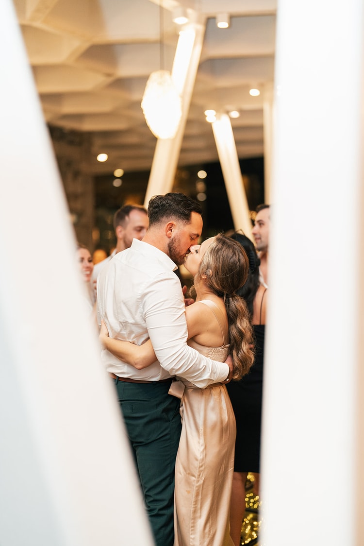 Bride and groom kiss on the dance floor while guests cheer and record with their phones under the pavilion lights.