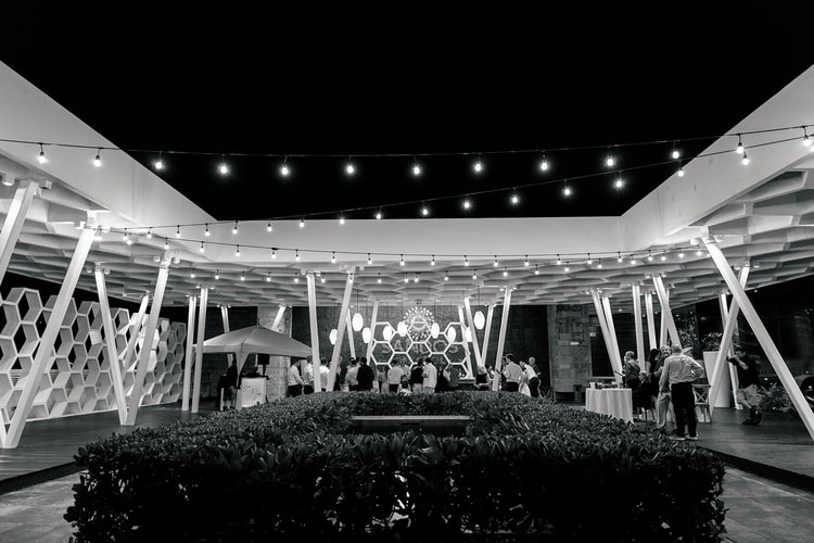 Wide view of the honeycomb-roofed reception pavilion strung with lights, guests gathered near the bar at night.