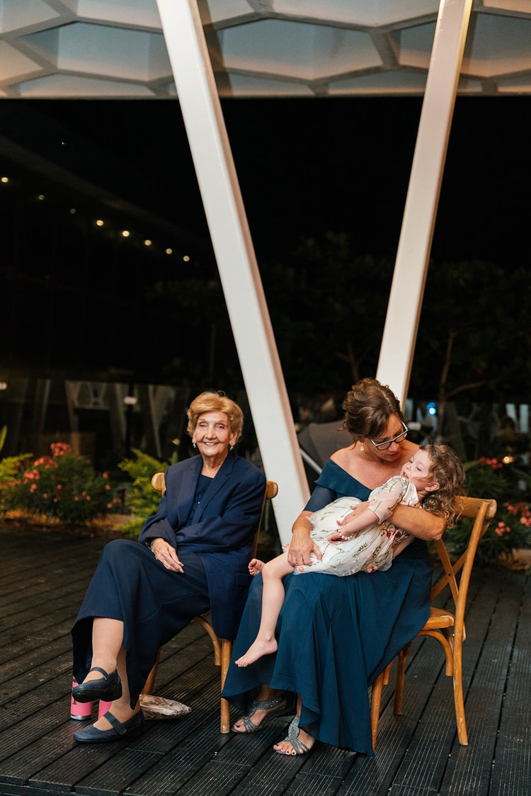 An elderly woman and a young girl in a floral dress rest together in chairs on the reception deck at night.