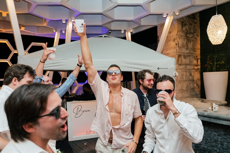 Groomsman raises a drink and cheers with friends near a pop-up bar tent bearing the groom's name at the reception.