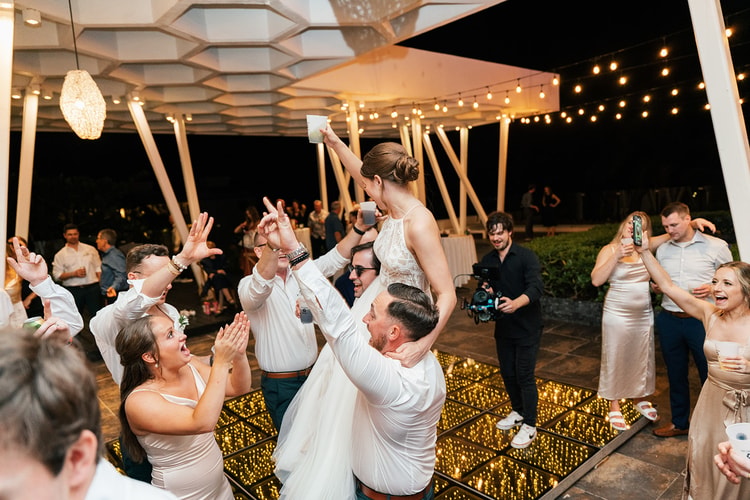 Groomsmen lift the bride on their shoulders as she raises a drink and guests cheer around the illuminated dance floor.