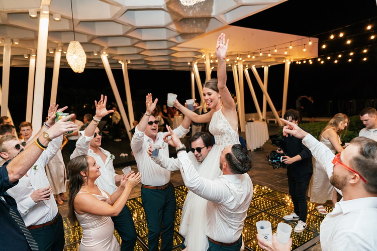 Bride raises her arms and cheers while dancing, surrounded by celebrating wedding guests under string lights.