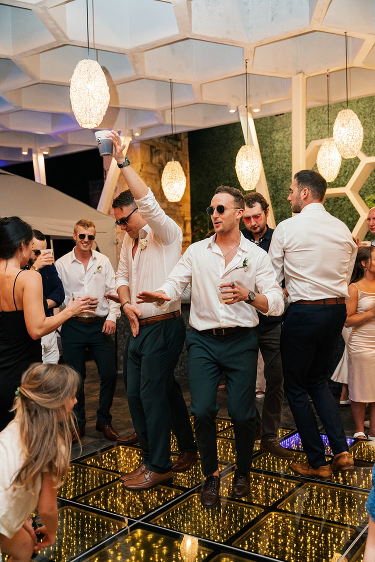 Groomsmen dance together on the glowing dance floor, one raising a drink overhead as a young girl watches nearby.