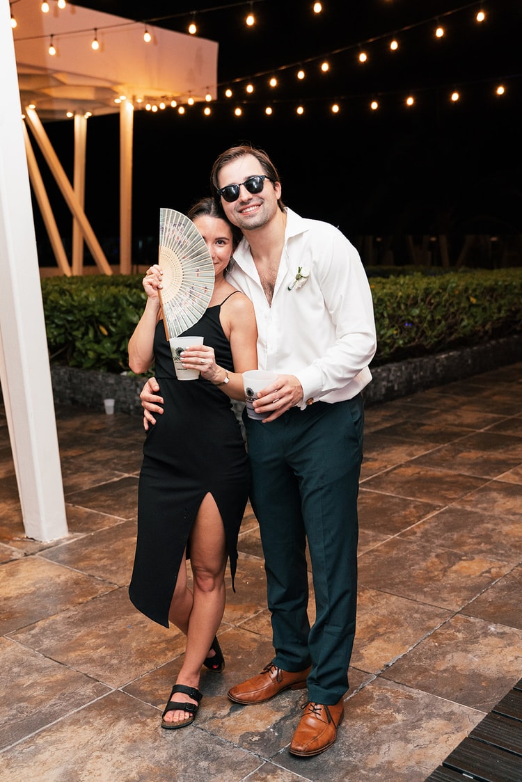 Wedding guest couple pose together at night, the woman peeking over a folding fan and both holding drinks.