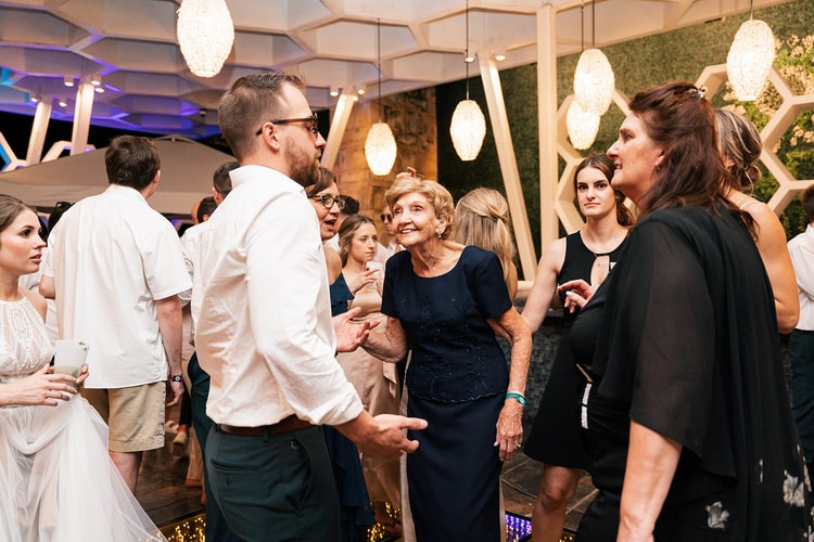 Groom chats with his smiling grandmother among a circle of wedding guests beneath hanging woven lanterns.