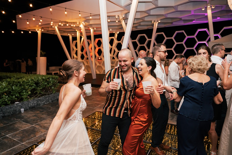 Bride laughs with two guests holding drinks near a honeycomb-patterned lounge structure lit in purple at the reception.