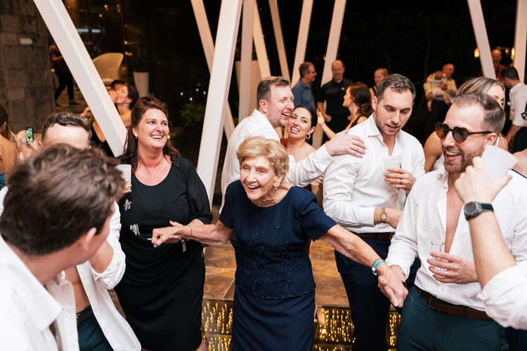 Elderly wedding guest in navy dances joyfully hand in hand with family members among a laughing crowd at the reception.