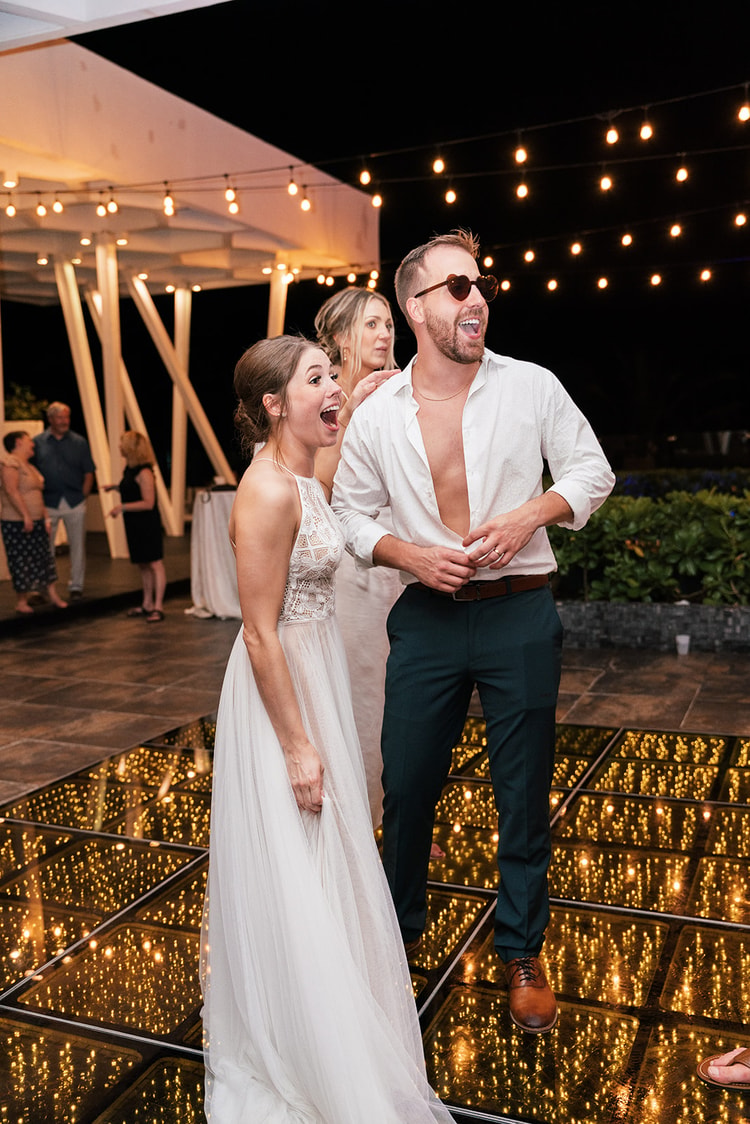 Bride and groom laugh together on the dance floor, the groom in heart-shaped sunglasses with his shirt unbuttoned.