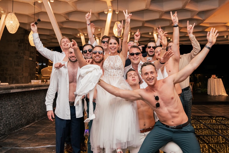 Bride and groom pose with a group of cheering groomsmen, several shirtless, throwing up hand signs under the pavilion.
