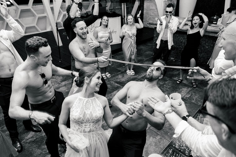 Bride laughs while playing a tie-tug game with a shirtless groomsman as guests clap and cheer around them.