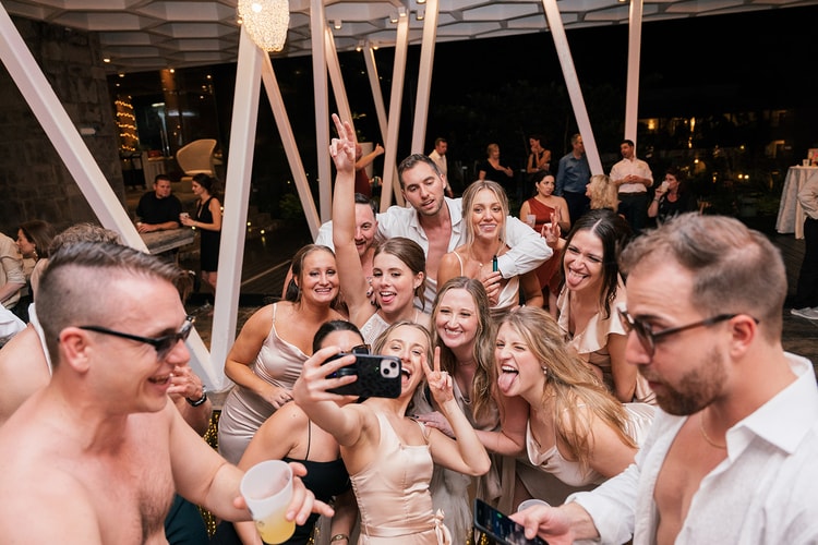 Bridesmaids and groomsmen crowd together for a phone selfie, laughing and posing on the dance floor at night.