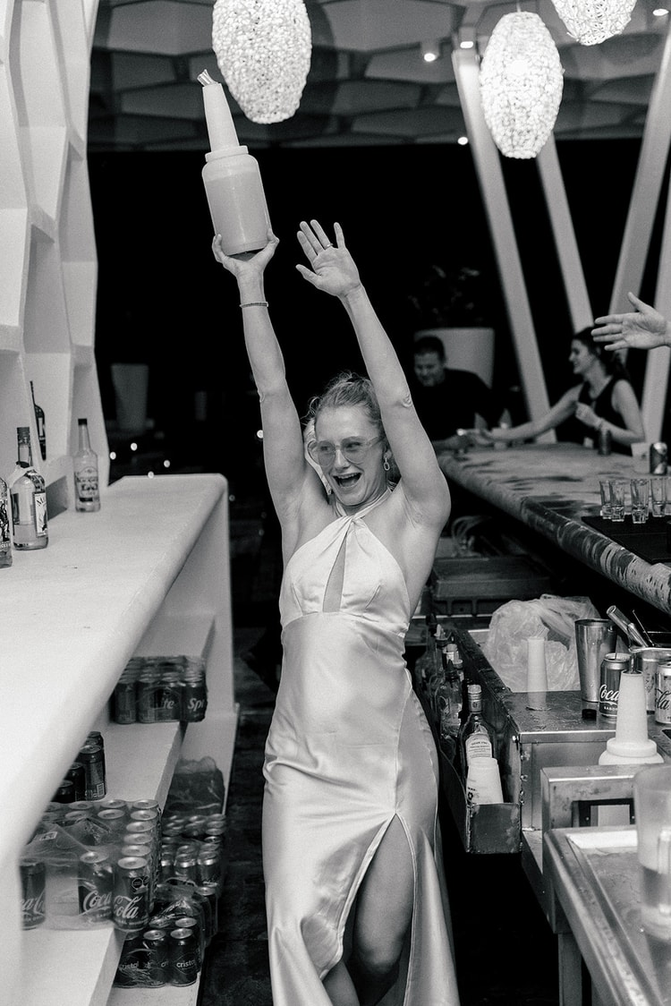 Bride raises a bottle triumphantly behind the bar, laughing among stacked soda cans and liquor bottles.