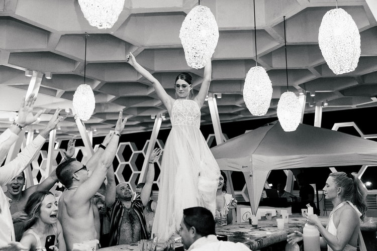 Bride stands triumphantly on a table, arms raised, as guests cheer beneath hanging woven pendant lights at night.