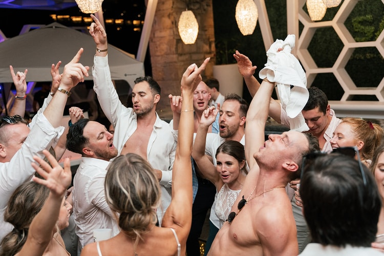 Guests dance in a tight circle, arms raised and singing along, some shirtless, beneath hexagonal pendant lights.
