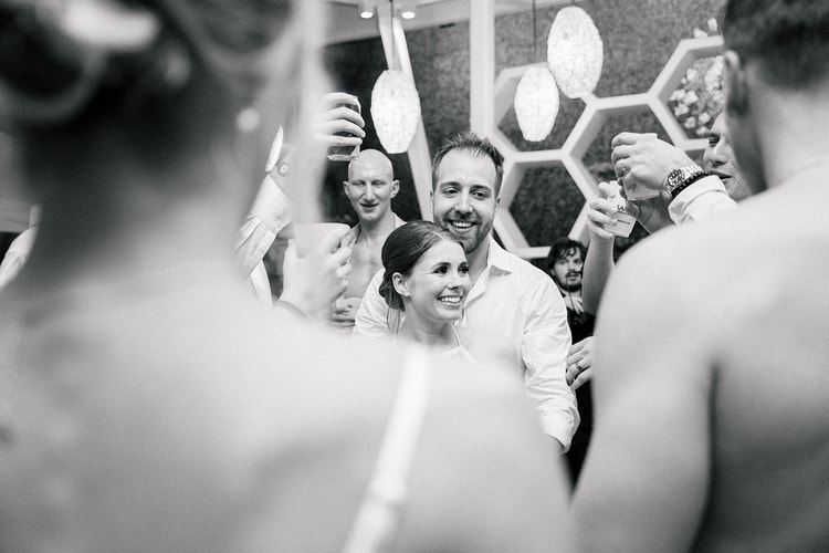 Bride and groom smile together as guests raise drinks around them in a close celebratory huddle at the reception.