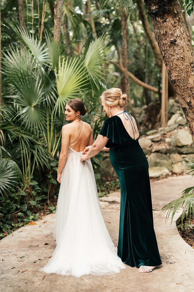 Bride in a spaghetti-strap gown smiles back over her shoulder as her mother in an emerald velvet dress fastens the back buttons amid jungle palms.
