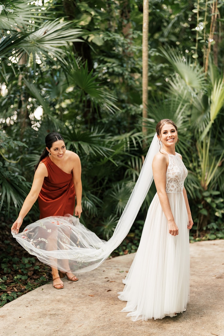 Bride smiles at a bridesmaid in a rust-colored dress who lifts and fans out the embroidered tulle train on a garden path.