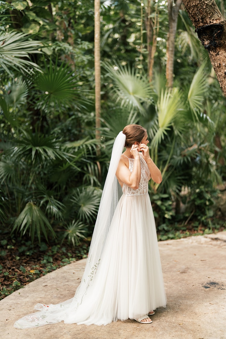 Bride in a flowing tulle gown and long veil adjusts her earring while standing on a stone path surrounded by palm fronds.