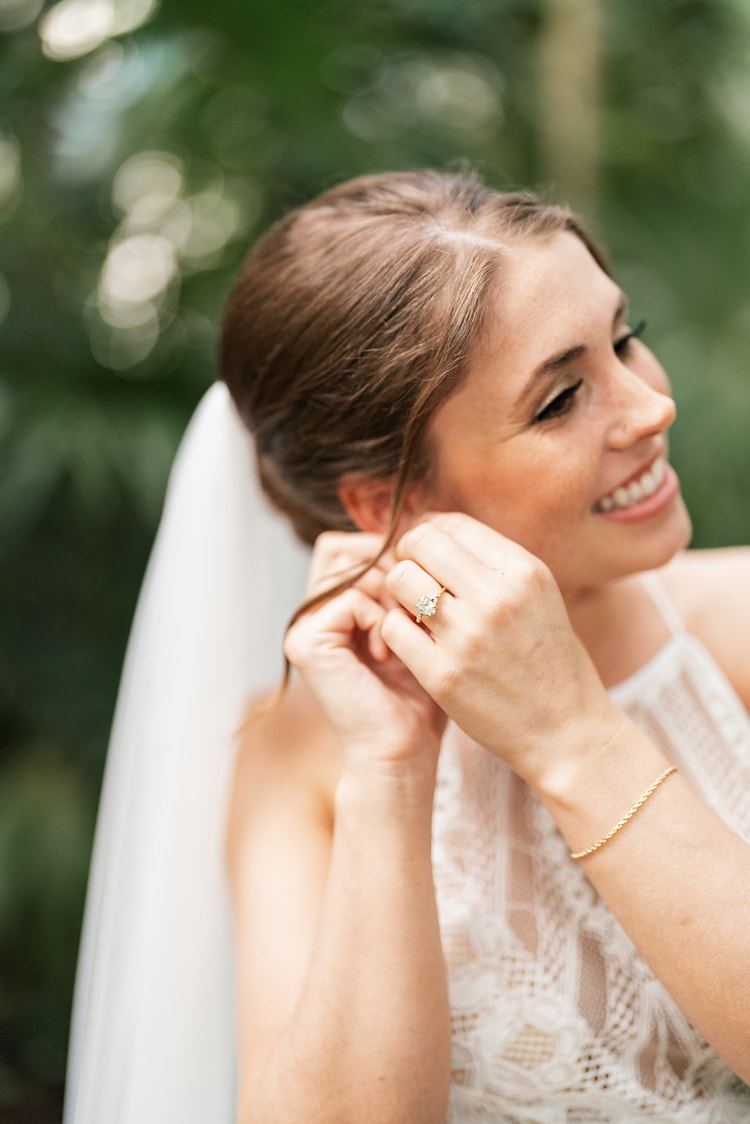 Bride smiling and reaching up to fasten an earring, her engagement ring visible, veil draped beside her in soft greenery light.