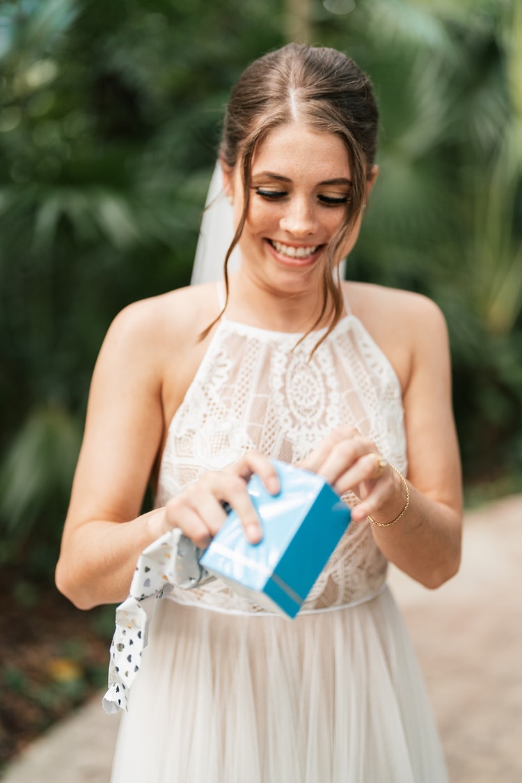 Bride in a lace halter gown and veil smiles while unwrapping a small gift box outdoors in dappled jungle light.