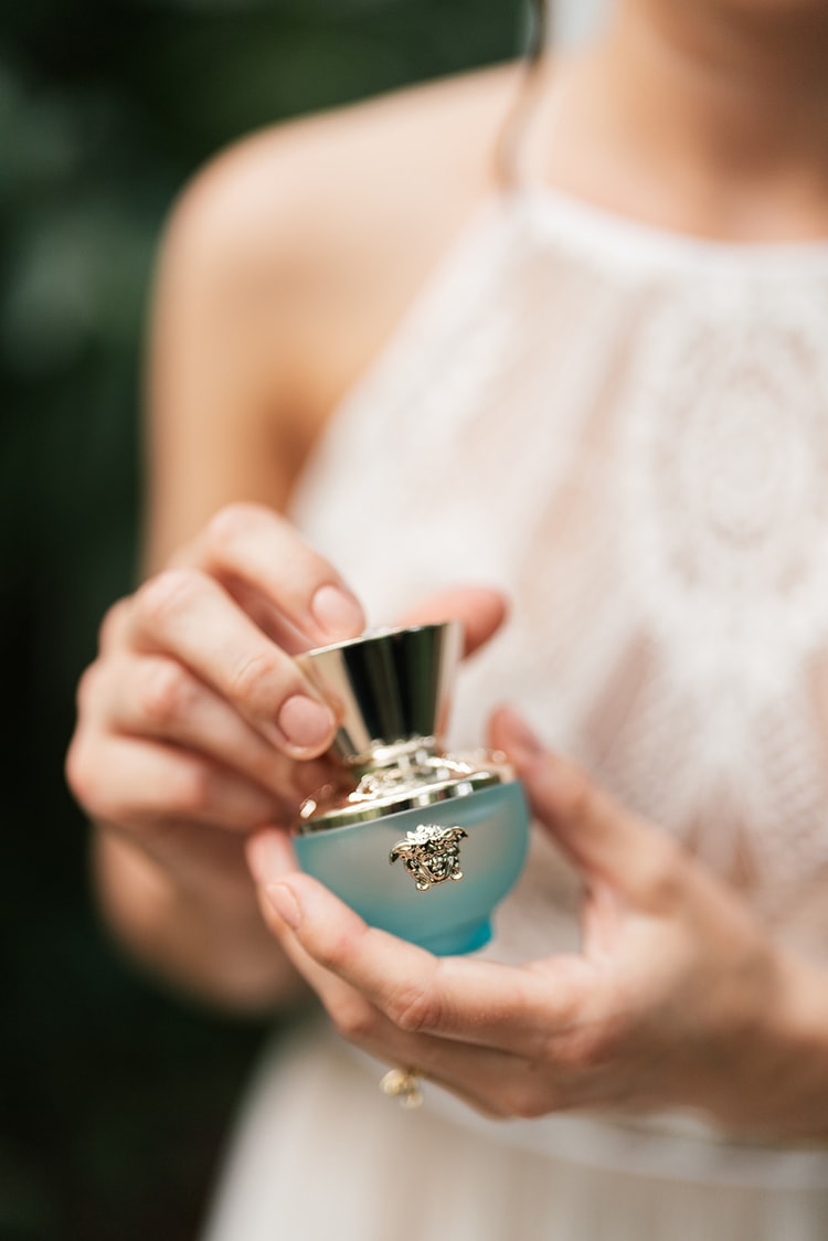 Close-up of a bride's hands opening a small blue perfume bottle with a gold Medusa emblem against her lace bodice.