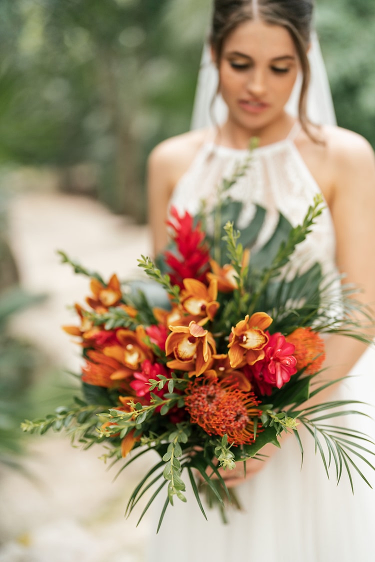 Close-up of a bride holding a bold tropical bouquet of orange orchids, red ginger, and pincushion protea against her lace gown.