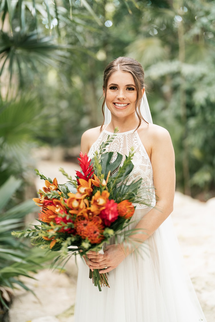 Smiling bride in a lace halter gown and veil holds a vibrant orange and red tropical bouquet in a jungle garden.
