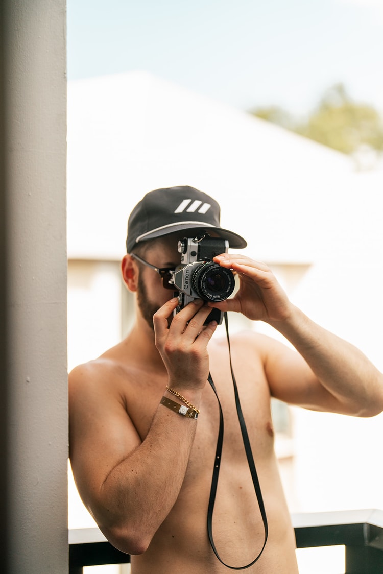 Shirtless groomsman wearing a cap and sunglasses photographs the scene with a vintage Canon film camera on a bright balcony.