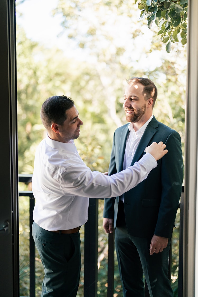 Groomsman in a white shirt helps a smiling groom in a teal suit adjust his jacket on a sunlit balcony surrounded by trees.