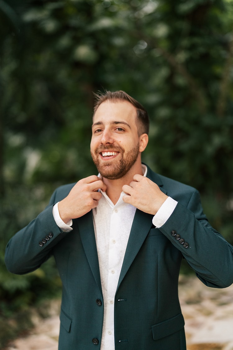Groom in a teal suit smiles while adjusting his white shirt collar, greenery blurred behind him.