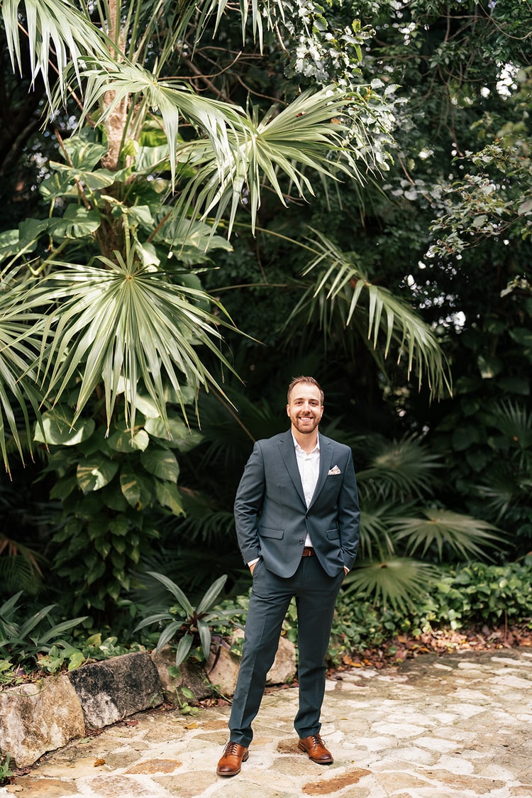 Groom in a teal suit with hands in his pockets stands smiling beneath a large palm tree on a stone pathway.