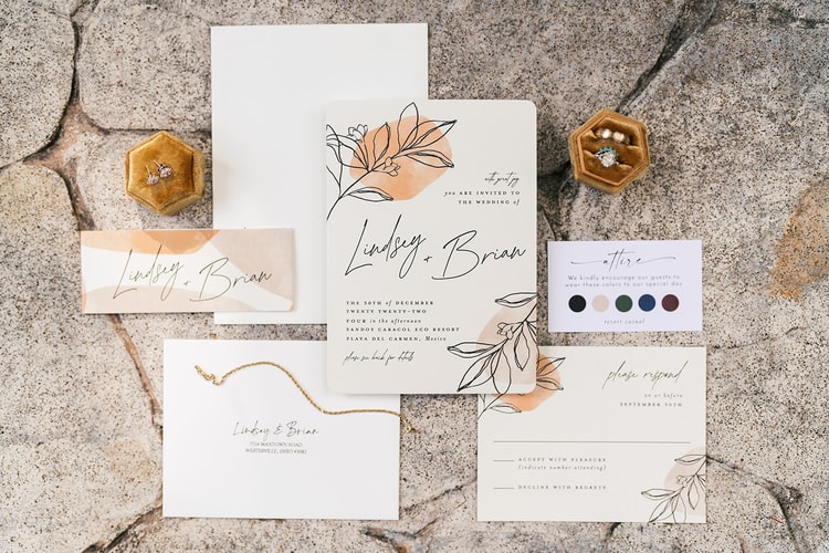 Wedding invitation suite with floral illustrations, gold rings, earrings, and a bracelet arranged on a stone surface for a Playa del Carmen, Mexico wedding.