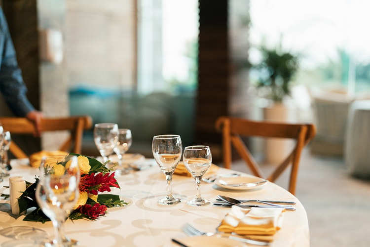 Reception table set with wine glasses, folded yellow napkins, and a small floral centerpiece of red and yellow blooms.
