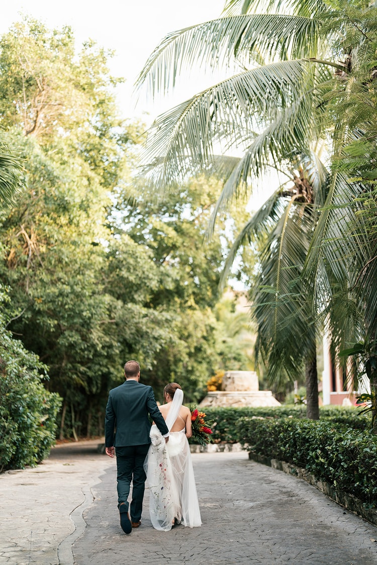 Bride and groom walk arm in arm down a stone path shaded by palm trees, seen from behind as they head into the resort grounds.