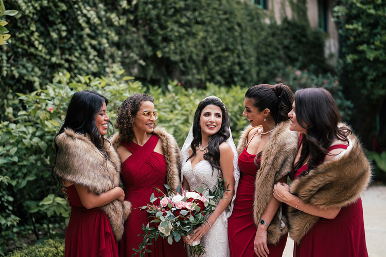 Bride laughs with bridesmaids in deep red dresses and fur stoles, holding a blush and burgundy bouquet, wedding photography