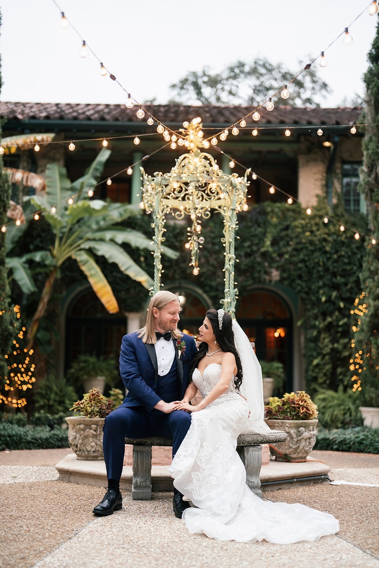Bride and groom on a stone bench beneath a gilded chandelier and cafe lights, Spanish-style estate behind, wedding photography