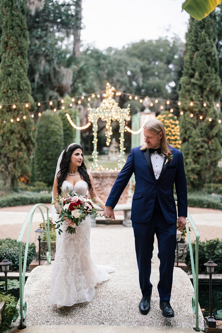 Bride and groom walk hand in hand across a garden bridge toward a glowing fountain in a cypress garden, wedding photography