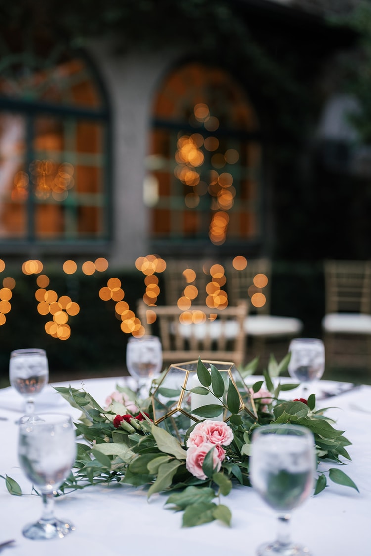 Reception centerpiece: geometric gold terrarium with pink roses and greenery, warm string lights glowing behind, wedding details