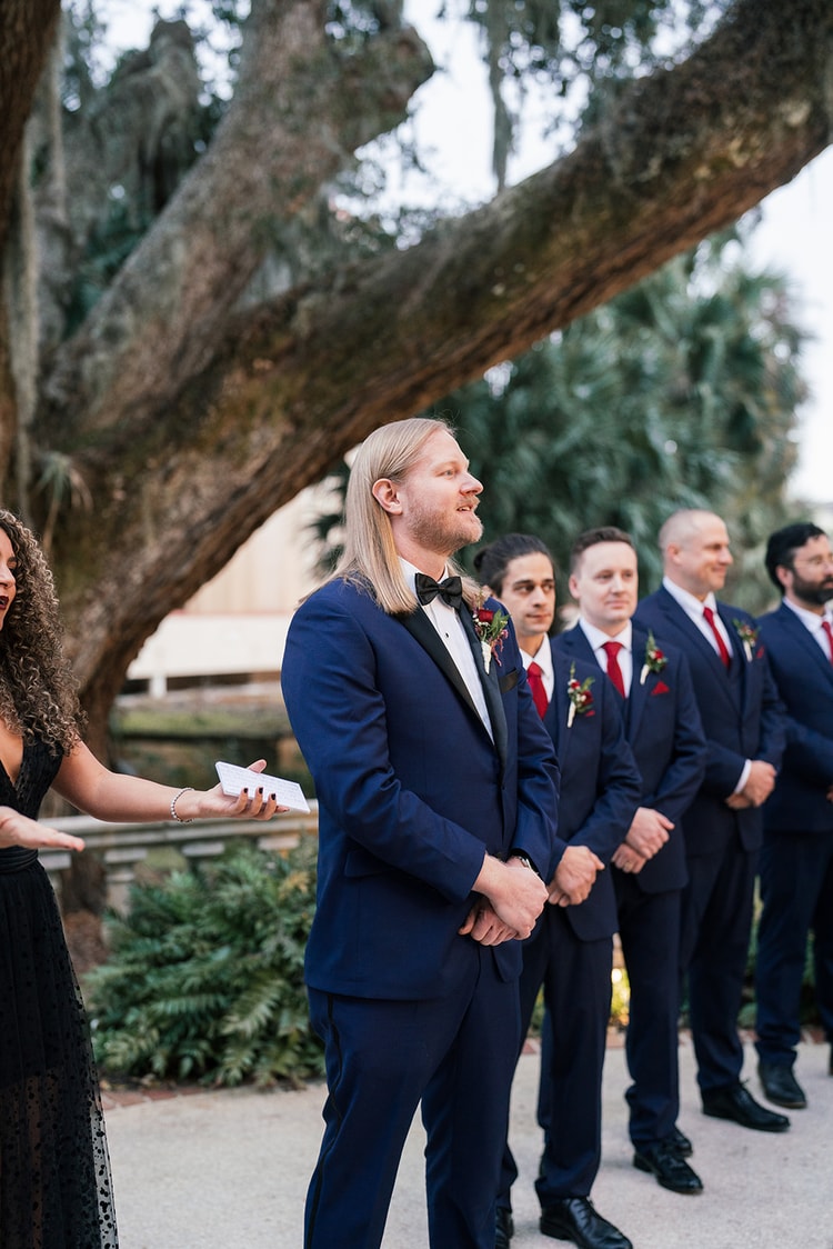 Groom watches the bride approach down the aisle with groomsmen lined up under a sprawling oak