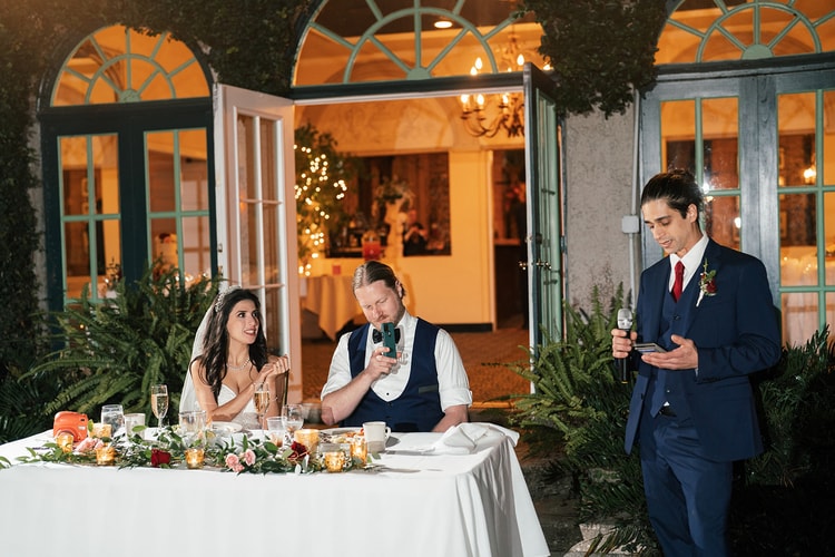 Bride raises a champagne glass laughing as the groom ducks and the best man delivers his toast outside