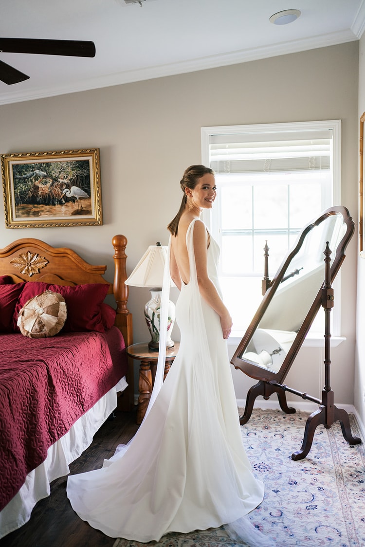 Bride in her open-back gown smiling over her shoulder at a full-length standing mirror in a warmly furnished bedroom.