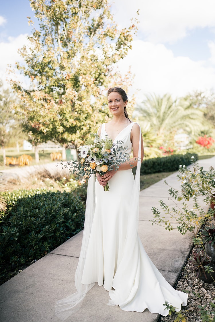 Bride in a sleek white gown with a flowing veil holding a wildflower bouquet, standing on a garden path surrounded by lush hedges and palms.
