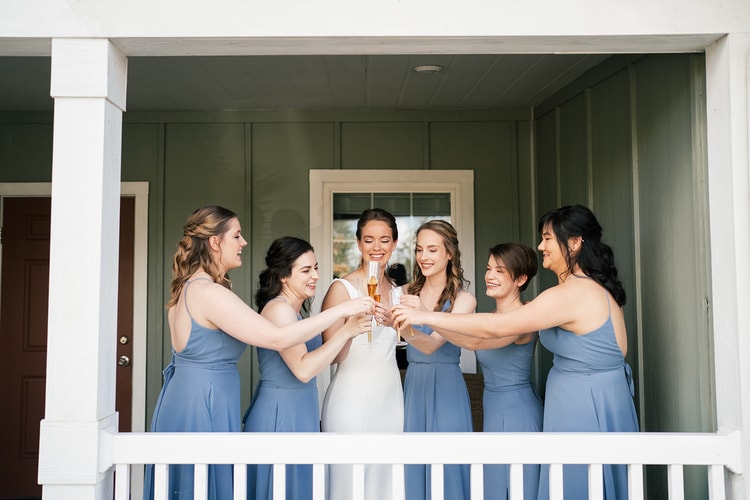 Bride and five bridesmaids in dusty blue gowns raising champagne flutes in a toast on a porch, laughing together.