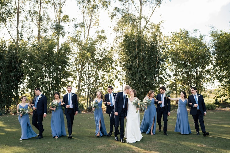 Wedding party walking together across a sunlit lawn with tall bamboo behind them, the couple at center kissing.
