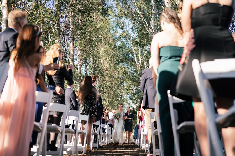 Bride walking down the outdoor aisle on a family member's arm, guests rising and turning to watch as she passes between rows of white chairs.